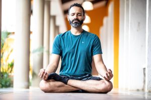 yoga-6950925_1280 Ein bärtiger Mann sitzt im Schneidersitz auf dem Boden eines Flurs, die Augen geschlossen, die Hände in meditativer Haltung auf die Knie gestützt, er trägt ein blaugrünes Hemd und schwarze Shorts.