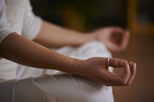 Eine Person in weißer Kleidung sitzt im Schneidersitz, die Hände ruhen in Meditationshaltung auf den Knien. Eine Hand ist sichtbar, deren Finger sich sanft berühren, und an einem Finger befindet sich ein silberner Ring.
