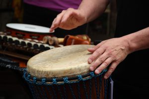 Eine Person spielt mit beiden Händen eine Djembe-Trommel. Im Hintergrund sind Perkussionsinstrumente wie ein Tamburin und ein Xylophon zu sehen.