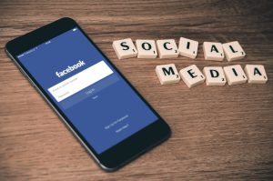 Ein Smartphone, das den Facebook-Anmeldebildschirm anzeigt, liegt auf einer Holzfläche neben Scrabble-Steinen, die so angeordnet sind, dass sie SOCIAL MEDIA buchstabieren.