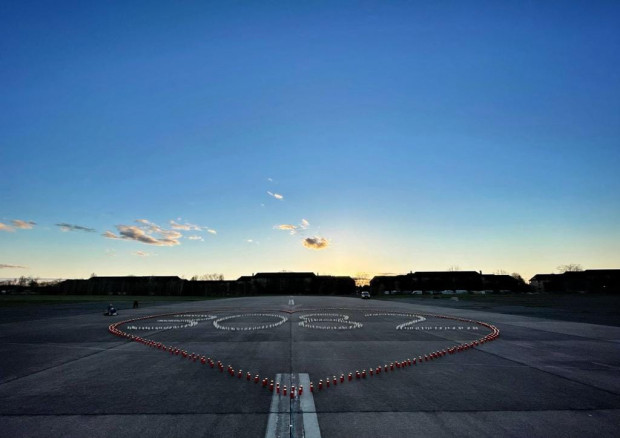 Bei Sonnenuntergang bilden auf einer breiten gepflasterten Fläche angeordnete Kerzen ein großes Herz um das Wort HOPE, während sich die Silhouetten der Gebäude vor dem farbenfrohen Himmel abzeichnen.