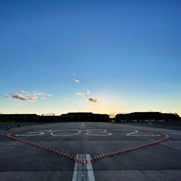 Bei Sonnenuntergang bilden auf einer breiten gepflasterten Fläche angeordnete Kerzen ein großes Herz um das Wort HOPE, während sich die Silhouetten der Gebäude vor dem farbenfrohen Himmel abzeichnen.