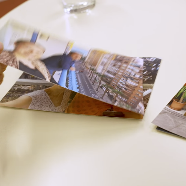 Eine Hand hält ein ausgedrucktes Foto über einem Tisch. Um ihn herum liegen weitere Fotos, die Menschen und Szenen im Freien zeigen. Daneben steht ein Glas Wasser.