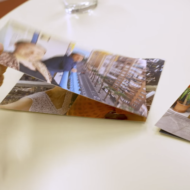 Eine Hand hält ein ausgedrucktes Foto über einem Tisch. Um ihn herum liegen weitere Fotos, die Menschen und Szenen im Freien zeigen. Daneben steht ein Glas Wasser.