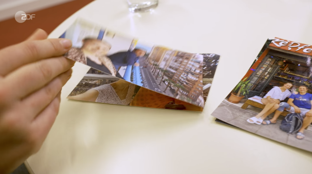 Eine Hand hält ein ausgedrucktes Foto über einem Tisch. Um ihn herum liegen weitere Fotos, die Menschen und Szenen im Freien zeigen. Daneben steht ein Glas Wasser.