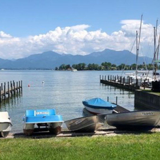 Kleine Boote liegen am Ufer neben einem Steg an einem ruhigen See, umgeben von Segelbooten, grünem Gras und fernen Bergen unter einem teilweise bewölkten Himmel.