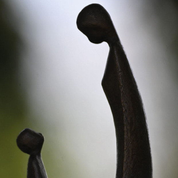 Eine Nahaufnahme einer glatten, abstrakten Skulptur, die zwei längliche Figuren zeigt, eine große und eine kleine, die sich vor einem unscharfen grün-grauen Hintergrund gegenüberstehen.