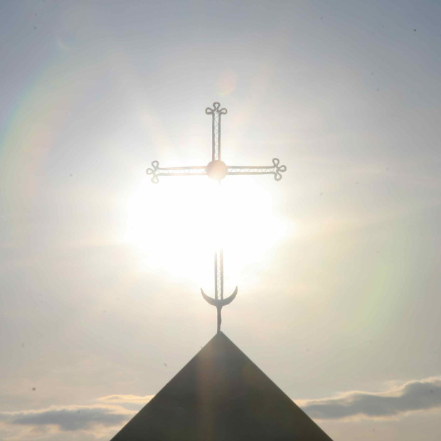Auf einem dreieckigen Dach steht ein von hellem Sonnenlicht beleuchtetes Metallkreuz, im Hintergrund ist ein bewölkter Himmel zu sehen.