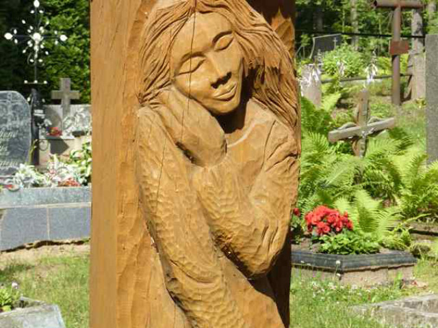 Holzskulptur einer gelassenen Frau mit geschlossenen Augen, die ihren Kopf auf ihre Hand stützt, in einen vertikalen Pfosten auf einem Friedhof im Freien geschnitzt, umgeben von Grabsteinen, Grünpflanzen und Blumen.