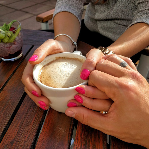 Zwei Personen sitzen an einem Holztisch. Eine hält eine Tasse Kaffee in der Hand, beide Hände sind mit rosa Nagellack lackiert, die andere hält sanft ihre Hand. Im Hintergrund stehen zwei Gläser mit geschichtetem Fruchtparfait.