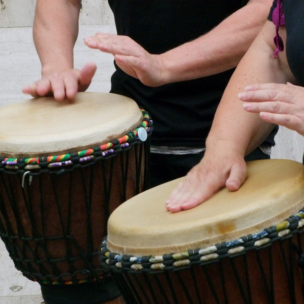 Zwei Personen spielen Handtrommeln und drücken ihre Handflächen auf die Trommelfelle. Die Trommeln haben bunte, geflochtene Ränder und einen dunklen Holzkorpus. Auf dem Bild sind nur die Arme und Hände der Spieler zu sehen.