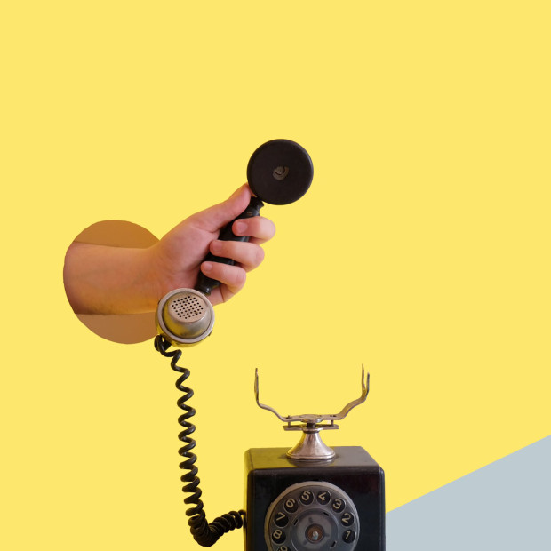 Eine Hand greift durch ein rundes Loch auf gelbem Hintergrund und hält den Hörer eines alten Wählscheibentelefons, das auf einer grauen Oberfläche liegt.