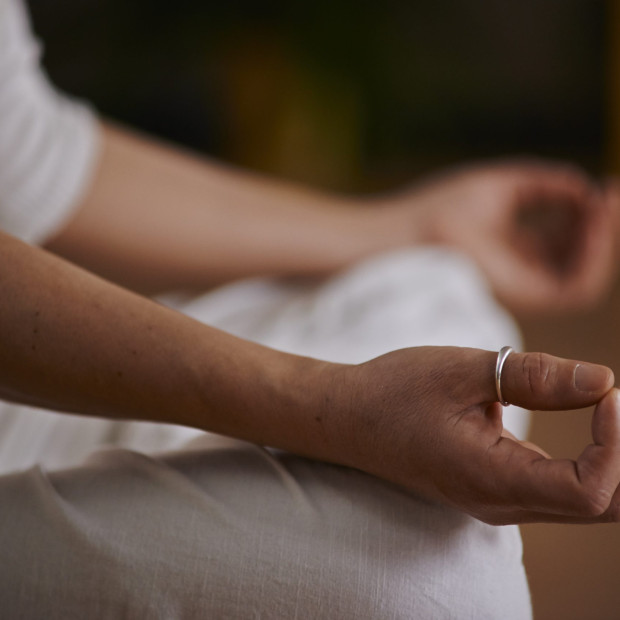 Eine Person in weißer Kleidung sitzt im Schneidersitz, die Hände ruhen in Meditationshaltung auf den Knien. Eine Hand ist sichtbar, deren Finger sich sanft berühren, und an einem Finger befindet sich ein silberner Ring.
