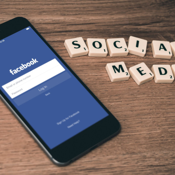 Ein Smartphone, das den Facebook-Anmeldebildschirm anzeigt, liegt auf einer Holzfläche neben Scrabble-Steinen, die so angeordnet sind, dass sie SOCIAL MEDIA buchstabieren.