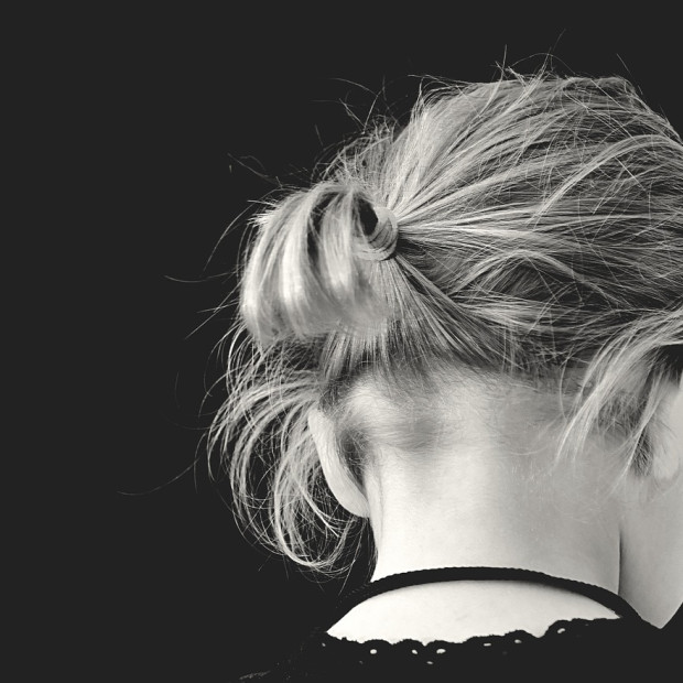 Ein Schwarzweißfoto einer Person mit hellem Haar, das zu einem unordentlichen Dutt gebunden ist, von hinten vor einem dunklen Hintergrund. Der Ausschnitt ihres Hemdes und ihr Nacken sind sichtbar.