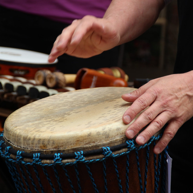 Eine Person spielt mit beiden Händen eine Djembe-Trommel. Im Hintergrund sind Perkussionsinstrumente wie ein Tamburin und ein Xylophon zu sehen.