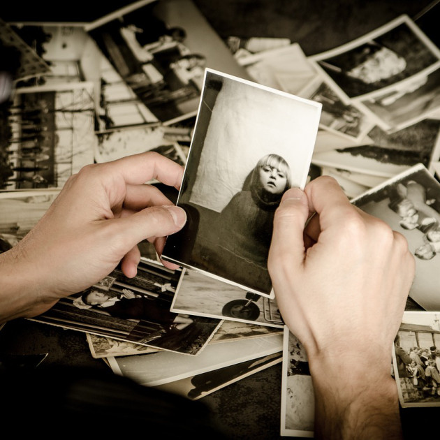 Eine Person hält ein Schwarzweißfoto eines Kindes, während sie eine verstreute Sammlung alter Fotos auf einem Tisch sortiert.