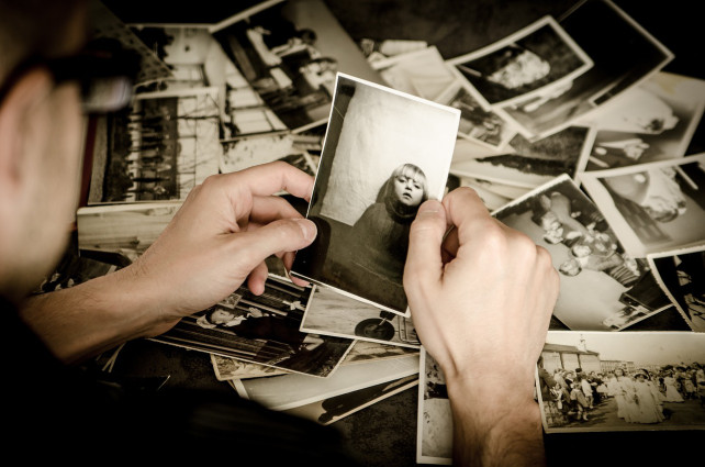 Eine Person hält ein Schwarzweißfoto eines Kindes, während sie eine verstreute Sammlung alter Fotos auf einem Tisch sortiert.