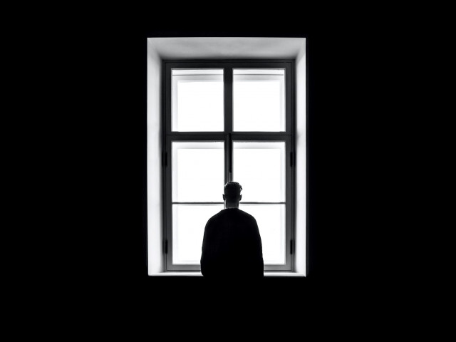 Rückansicht eines Mannes, der aaus einem dunklen Raum aus dem Fenster schaut