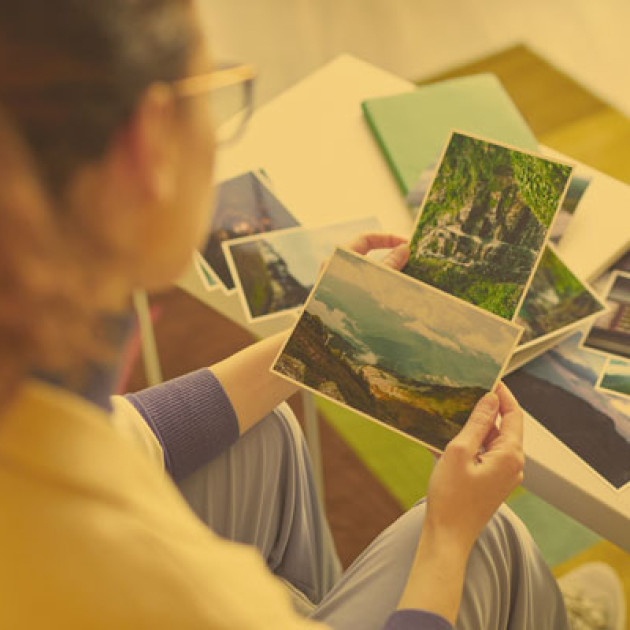 Eine Person mit langen Haaren und Brille sitzt an einem Tisch und betrachtet mehrere Landschaftsfotos. Weitere Fotos und Notizbücher sind auf dem Tisch ausgebreitet. Die Szene scheint in einem Innenraum mit weicher Beleuchtung zu spielen.
